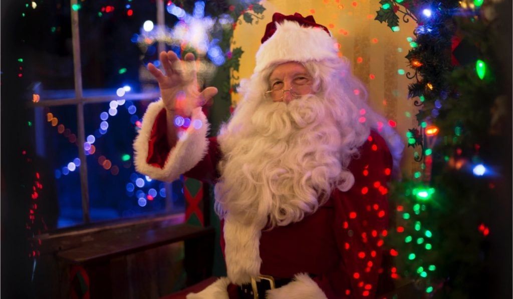 Santa Claus sitting down and waving. There are Christmas lights around him