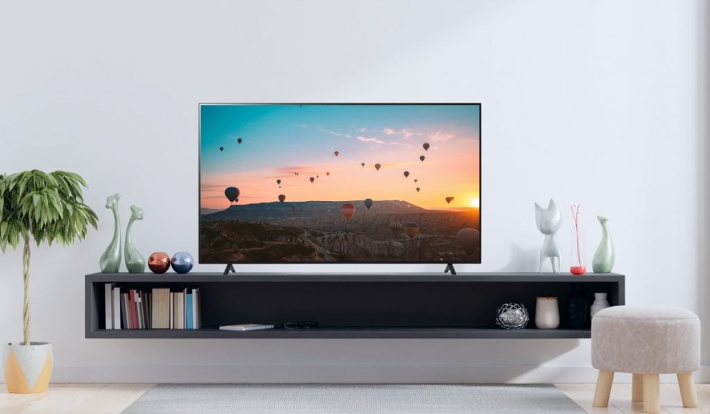 Een Smart TV met een foto van een zonsopgang boven een vlakte. de tv staat op een lage plank. Er is een plant en een stoel aan weerszijden van de plank