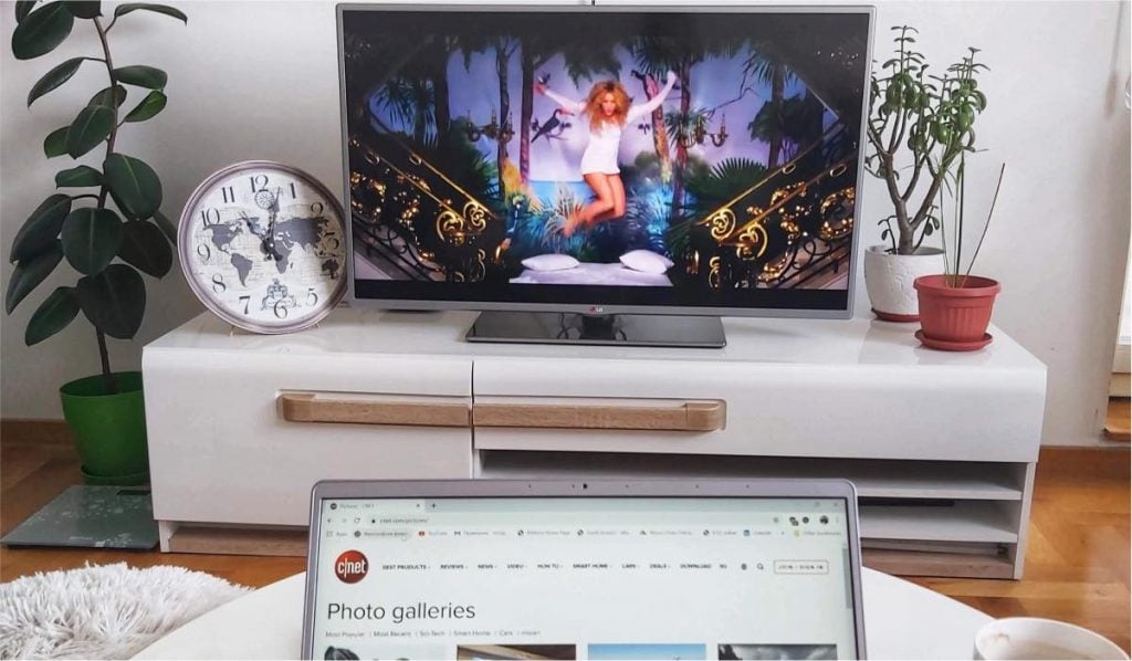 a top half of laptop screen at the bottom of the image. A TV on a white TV stand in a living room with a plant and a large clock on either sides of the TV.