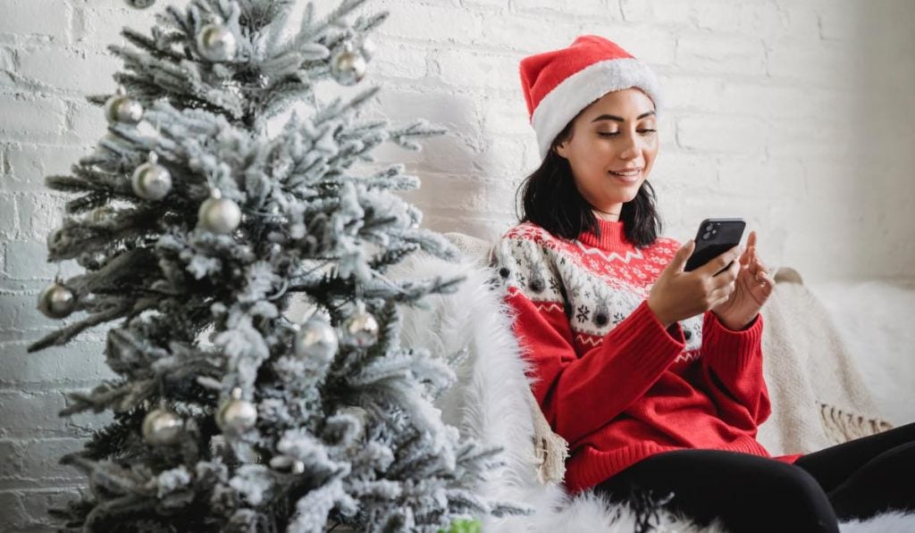 a woman in a christmas sweater and a santa claus hat sitting on a couch scrolling her smartphone. There is a decorated christmas tree to her side
