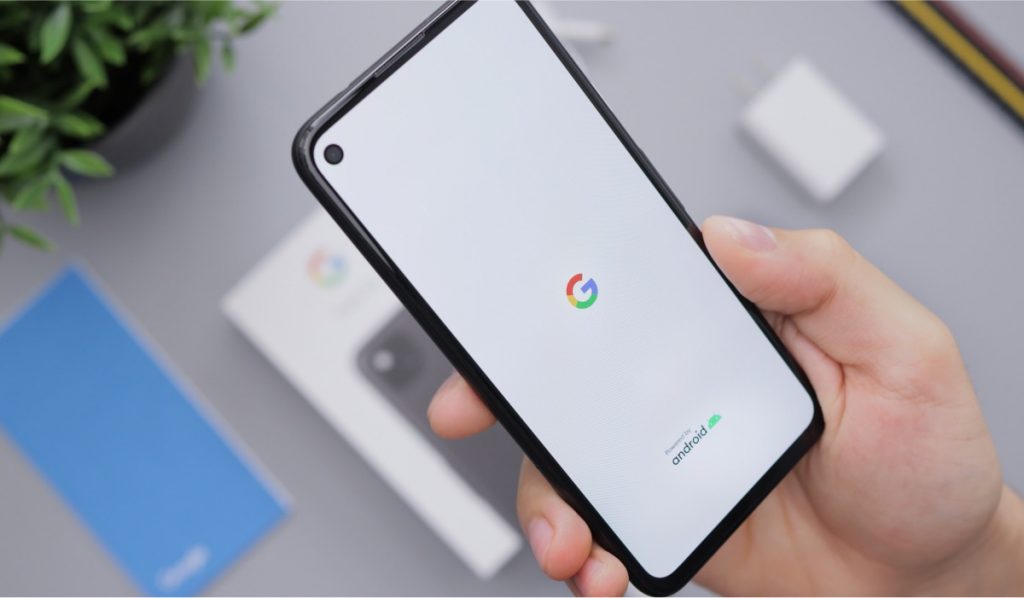 A hand is holding a Google Android smartphone. The smartphone is booting up. Blurred in the background is the smartphone box, a charging cube, a manual and a potted plant.