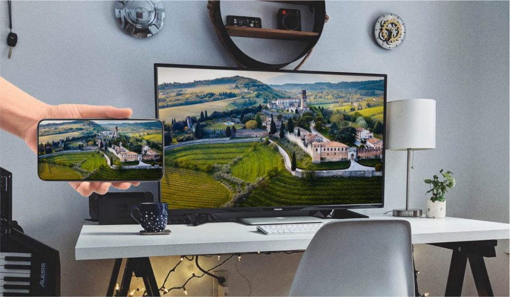 Une main tient un smartphone qui projette une image de drone d'un paysage de vieille ville sur une Smart TV. La Smart TV est posée sur un bureau blanc.