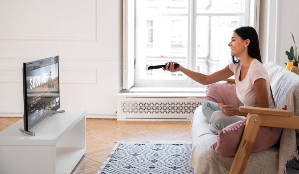 A woman is sitting cross-legged on a wood-framed couch. The is pointing a remote control at a Smart TV that is displaying a streaming service interface with 'Survivor' show page on.