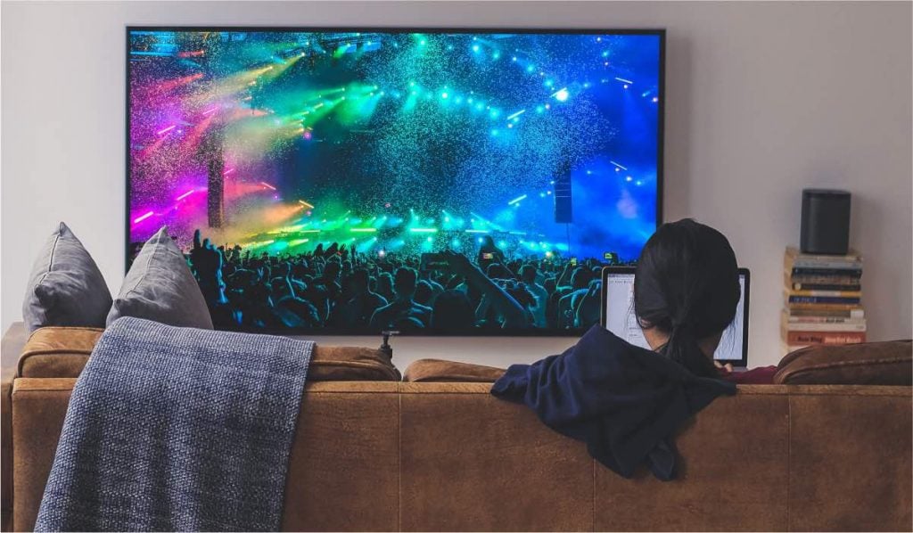 Une femme est assise sur un canapé et utilise un ordinateur portable. Une couverture est accrochée au siège du canapé. Une télévision est posée sur un meuble. La télévision affiche l'image d'un grand concert avec un spectacle de lumières.