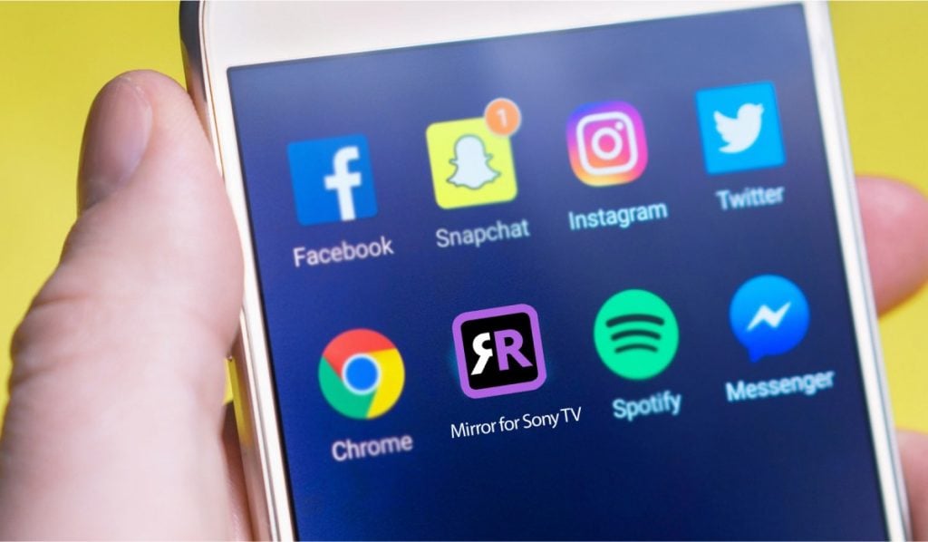 A hand holding an Android smartphone with several app icons on the screen - Facebook, Snapchat, Twitter, Instagram, Chrome, Mirror for Sony tV, spotify and messenger