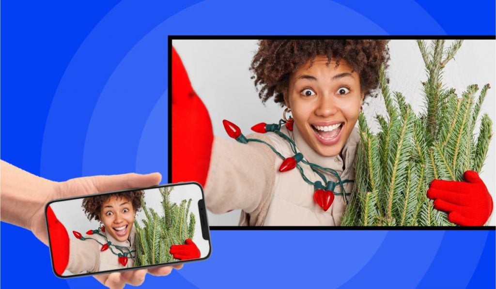 A hand holding a smartphone. The smartphone is mirroring an image of a woman taking a selfie while holding an undecorated Christmas tree and with Christmas lights around her neck to a Smart TV.