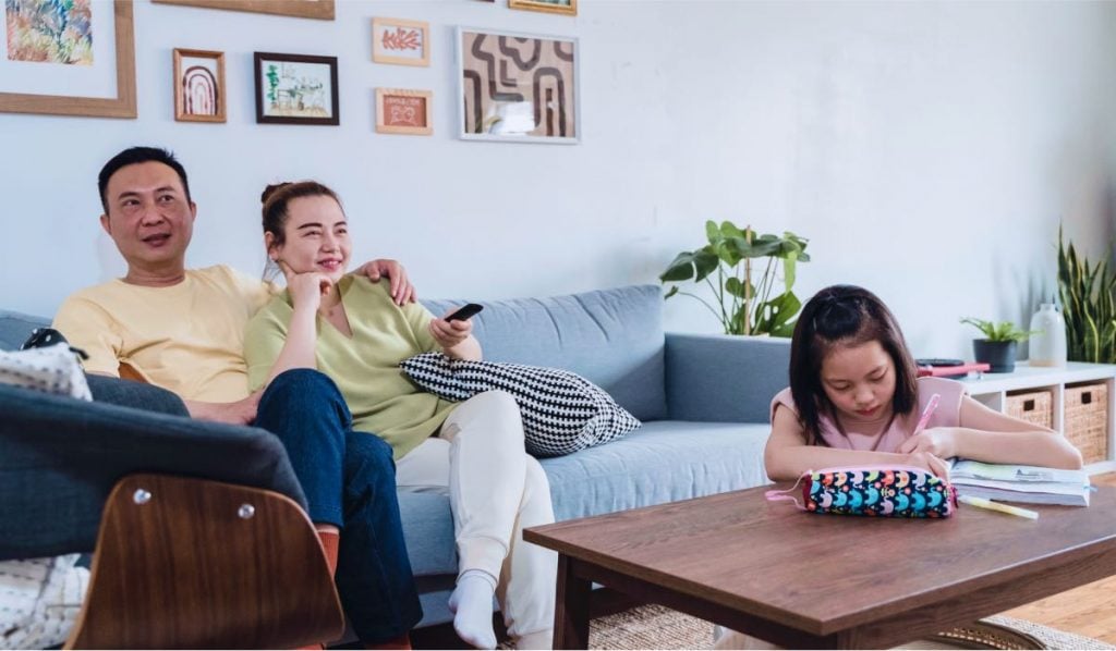 A man and a woman sit on a couch in an embrace. The woman is holding a remote. There's a child sitting by a coffee table with a coloring book.