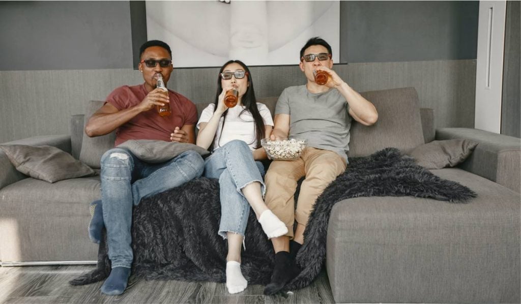 Two men and a woman sit on a couch. They are wearing sunglasses. Each of them is drinking a beer out of a glass bottle. The man on the right is holding a bowl of popcorn.