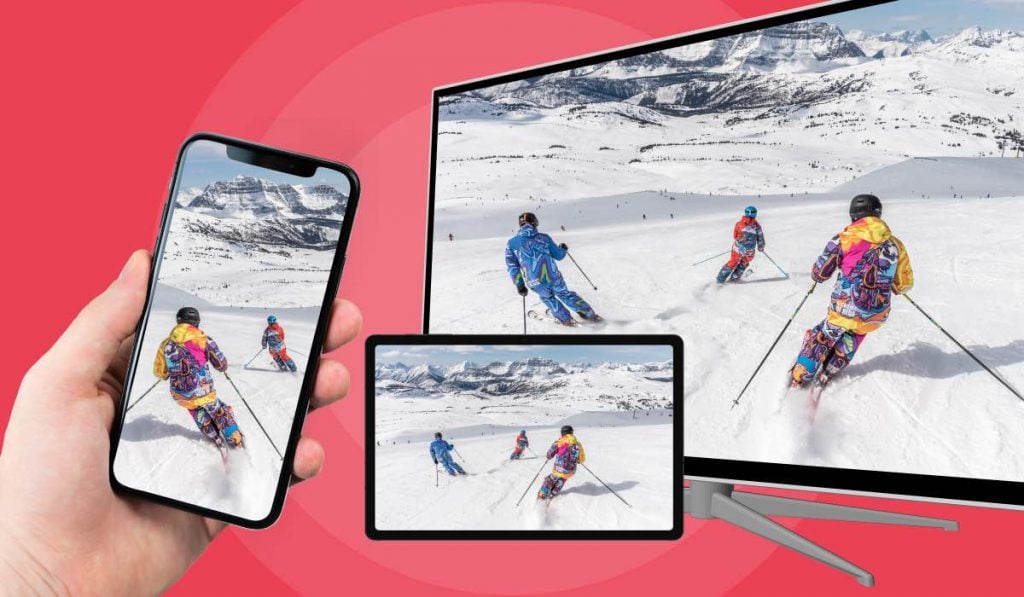 A hand holding an iPhone, a tablet and a Smart TV. Each device displays the same image of people skiing in a winter landscape. The background of the image is red.