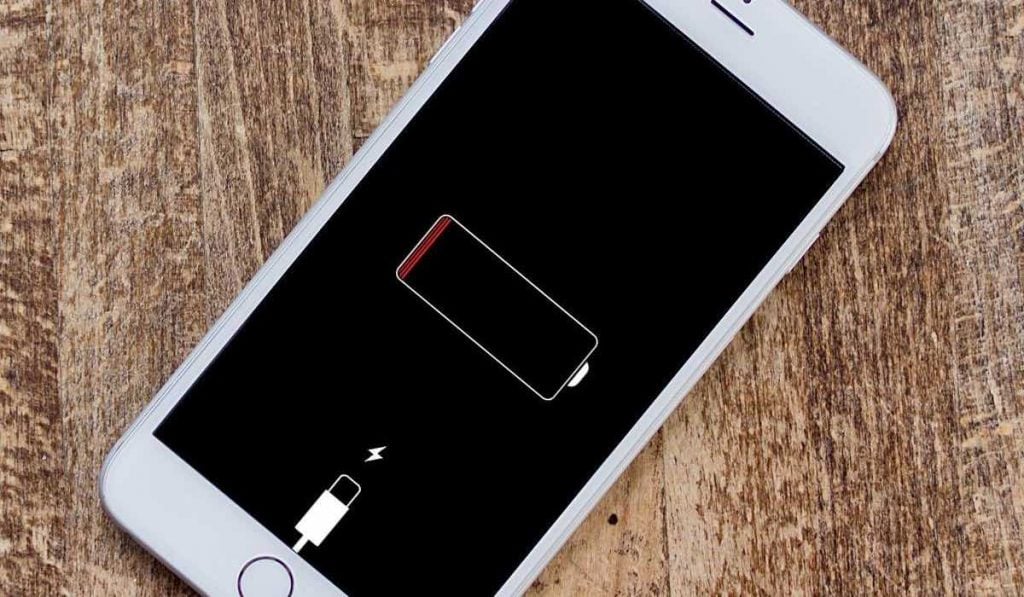 A white iPhone on a wooden surface. The screen shows a notification that the battery is empty and the iPhone has to be plugged in to charge.