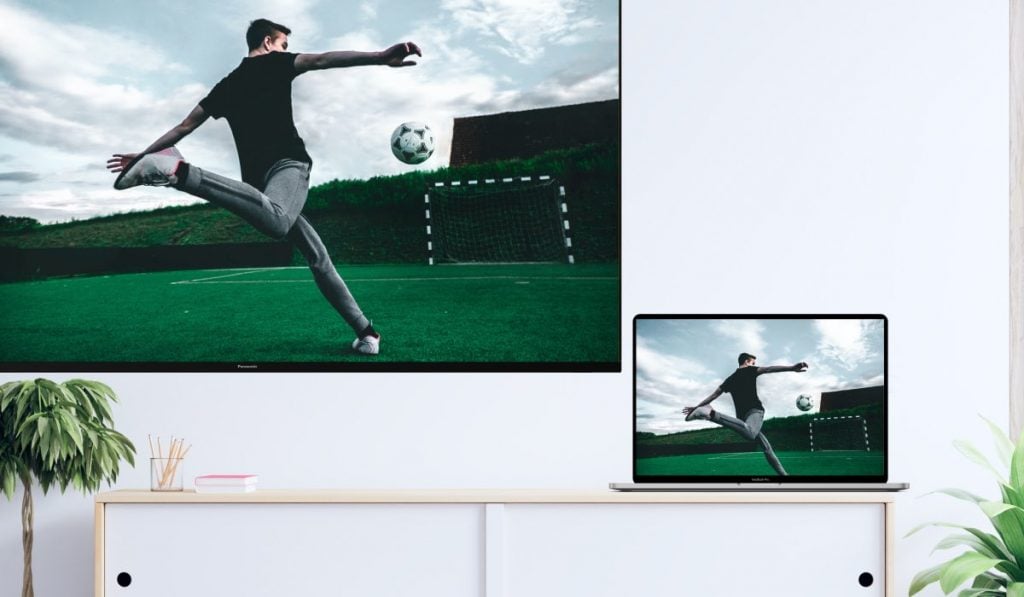 A MacBook and a Smart TV displaying the same image of a football player kicking a ball. The TV is hanging on a wall and the MacBook is resting on a piece of furniture.