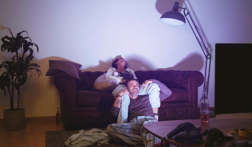 A man and a woman sit together on a couch while laughing. They are illuminated by light coming from a TV. There is a bottle of beer on a coffee table in front of them.