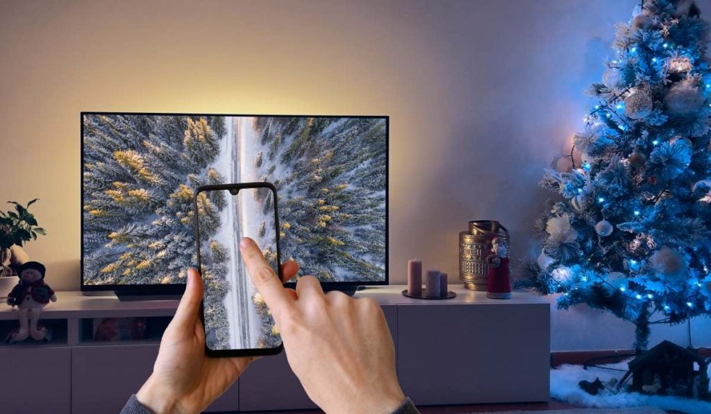 A hand holding a smartphone. The smartphone displays a drone shot of a road in a forest with a car on it. The image is mirrored to a Smart TV. The TV stands on a TV stand with a decorated Christmas tree on the right side of the image.