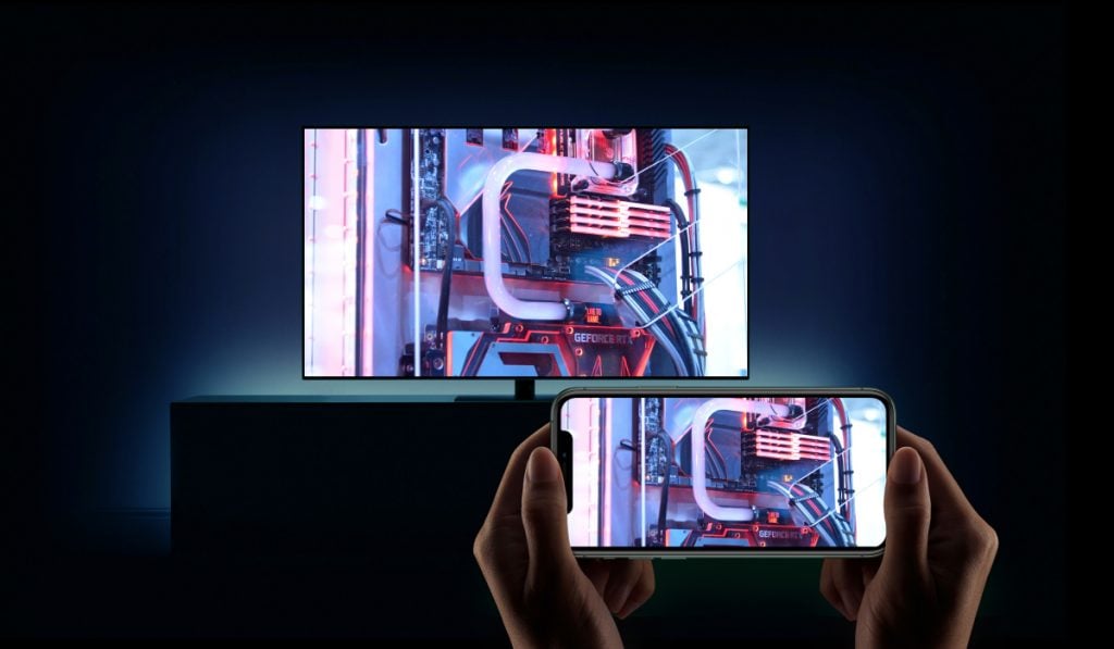 Two hands holding a smartphone horizontally. The smartphone is mirroring an image of pipes to a TV. The room is dark.