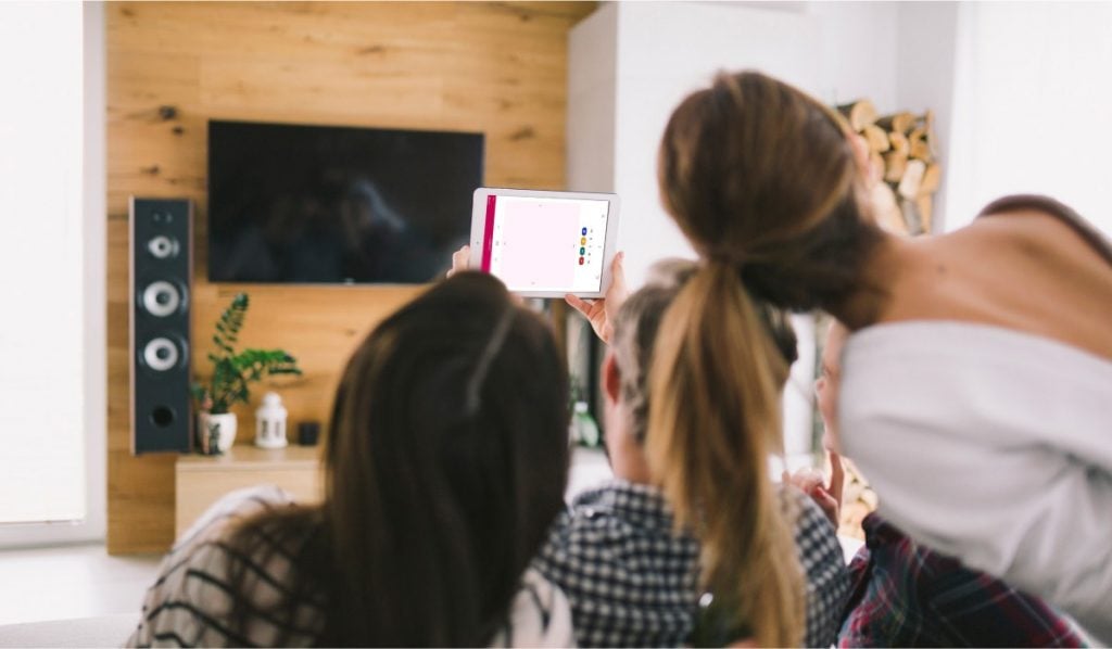 Three women using the LG TV remote app by MeisterApps. There is a Smart TV on the wall in front of them. There are two surround sound system speakers around the TV