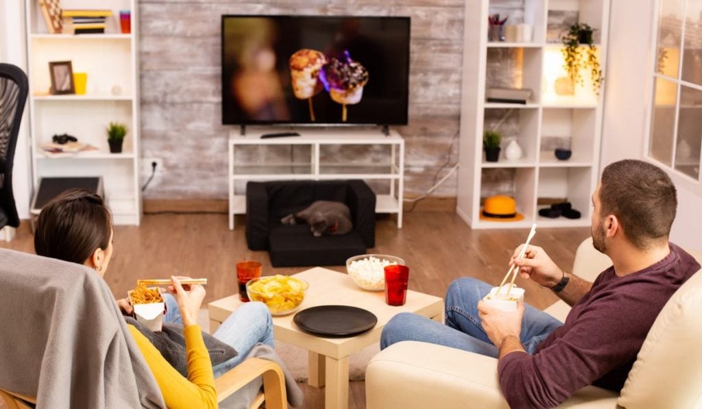 Two people sit in two comfortable chairs and watch TV. There are snacks on the table in front of them and some furniture around the TV.