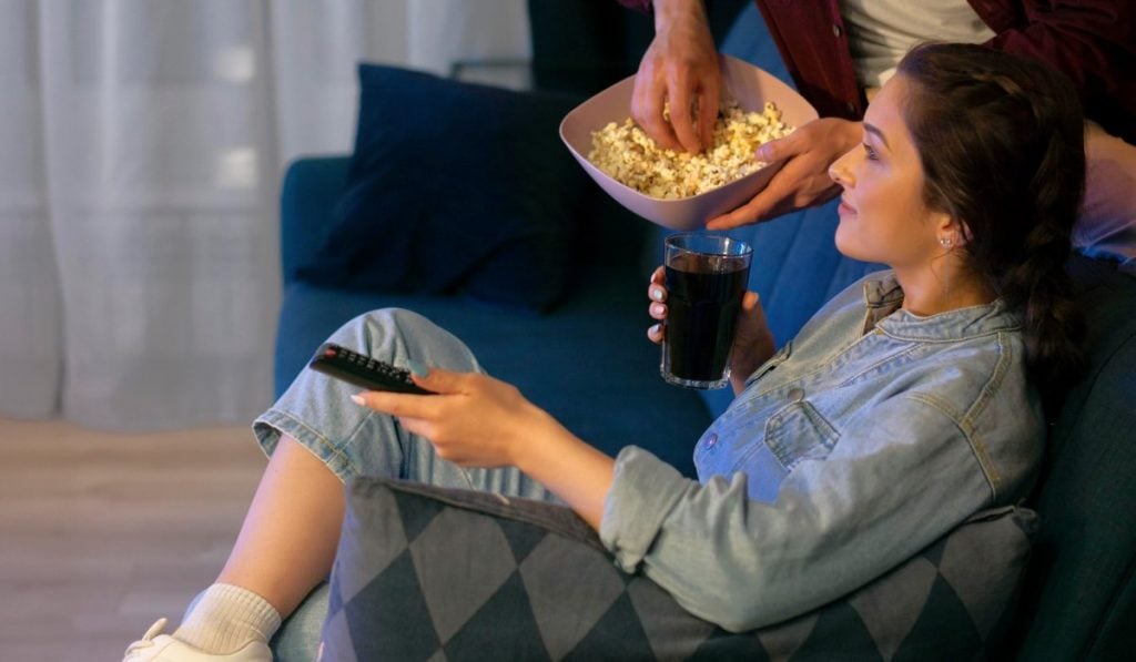 A woman is sitting on a couch with a remote control in her hand. There's a hand in a bowl of popcorn next to her.