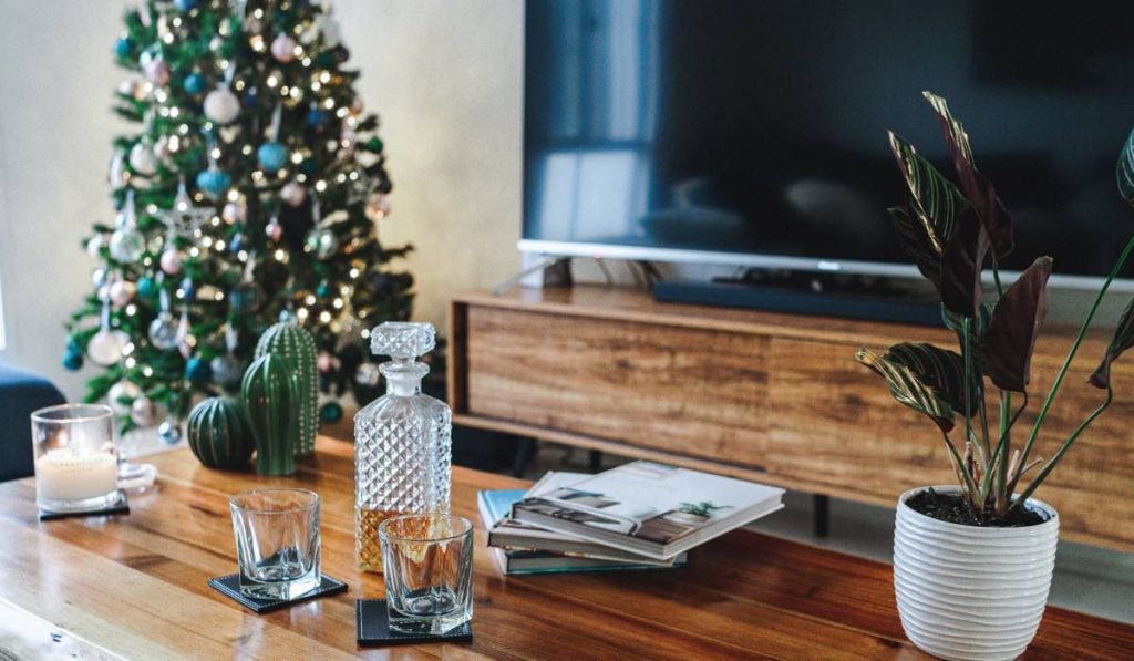 A wooden table with a carafe of dark liquor, two whisky glasses and a potted plant. There is a Smart TV in the background, as well as a decorated Christmas tree.