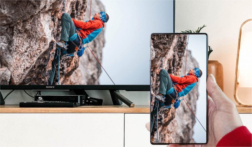 An Android smartphone casting an image of a person hiking among rocks to a Sony TV that is standing on a white TV stand.