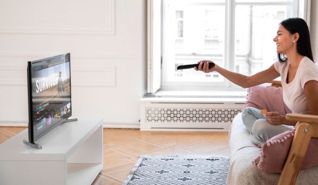 A woman is sitting on a couch with her legs crossed. She is smiling and pointing a remote control at a TV in front of her. The TV shows a streaming service interface for Survivor.