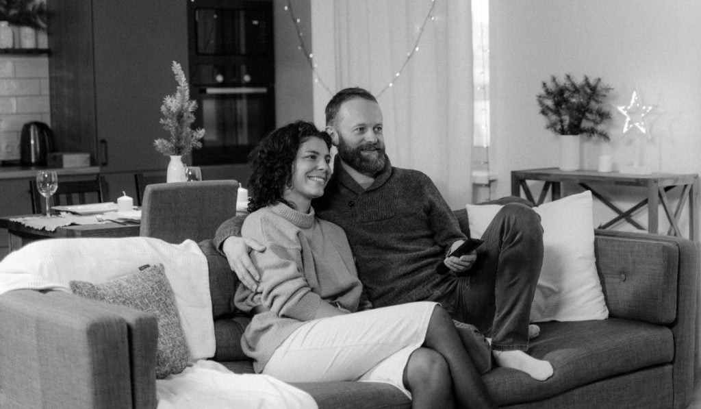 A black and white image of a man and a woman sitting on a couch in an embrace. They are located in a living room with some furniture arounnd them.