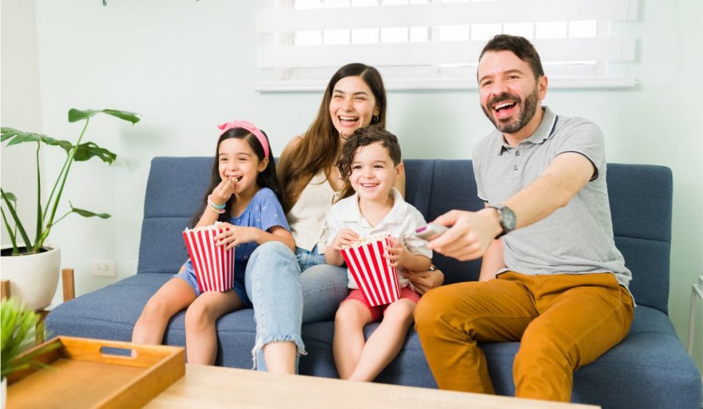 A family - man, a woman, and two kids eat popcorn out of cimena-style containers while sitting on a couch. The father is holding a remote control.
