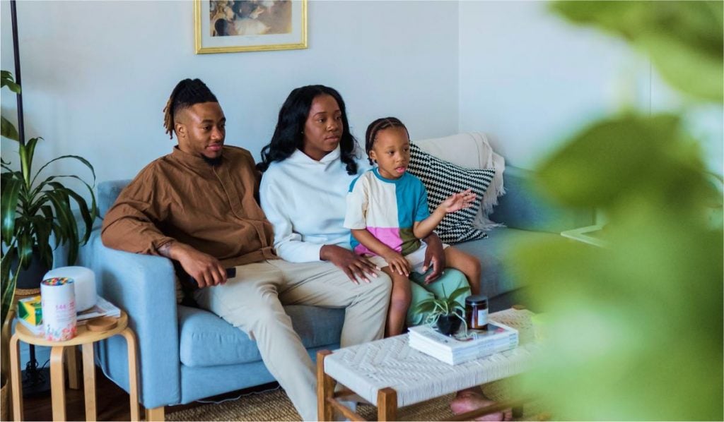 A family or three, a child, a mother and a father is sitting on a couch watching TV.