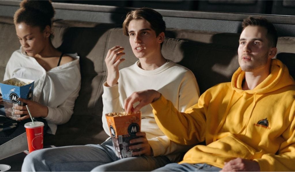 Three people - one woman and two men are sitting on a couch eating snacks.