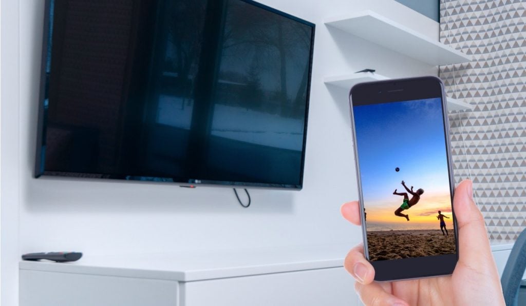 A hand holding a smartphone with an image of a person kicking a ball in mid-air during a sunset. There's an LG Smart TV hanging on a wall with white shelves around it. The TV has a black screen.