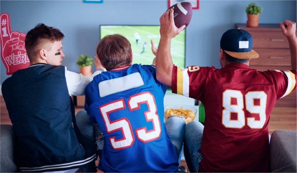 Three men sit on the couch. Two of the men have numbered football t-shirts on. They are watching sports on TV and cheering.
