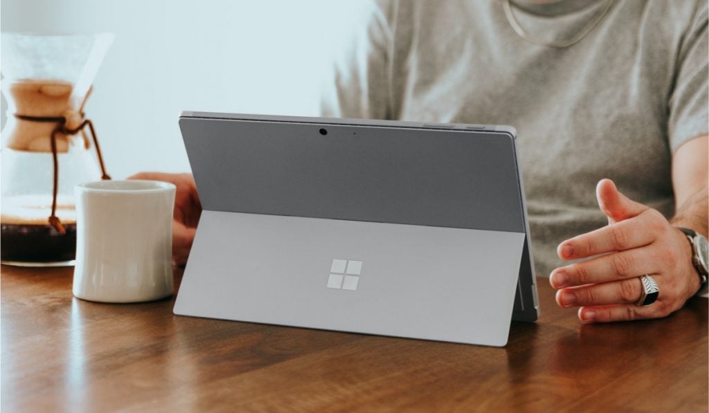 A person sitting at a table with a coffee mug, a chemex and a Microsoft tablet.