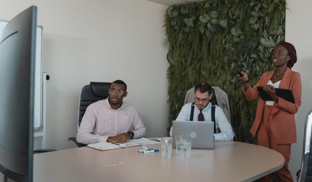 Two men sit at a large table and a woman is standing with a tablet. They are in a meeting room, looking at a Smart TV. There's a plant wall behind them