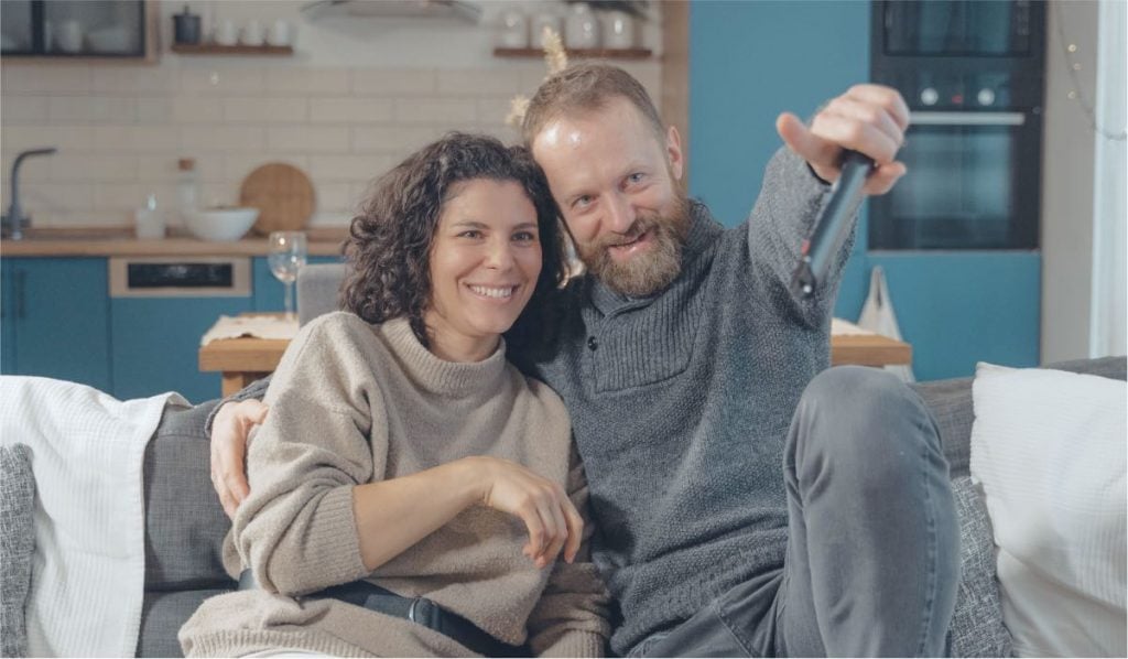 A man and a woman sit on the couch. They are smiling. The man is holding a remote control. In the background there are some kitchen appliances.