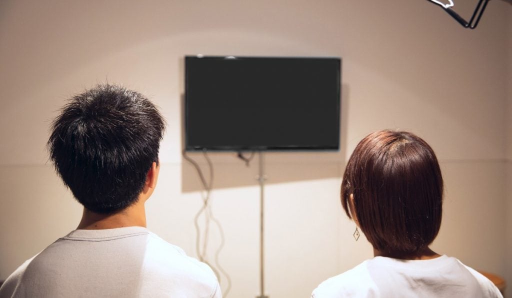 Two people watching a small Smart TV hanging on a wall with cables plugged in.