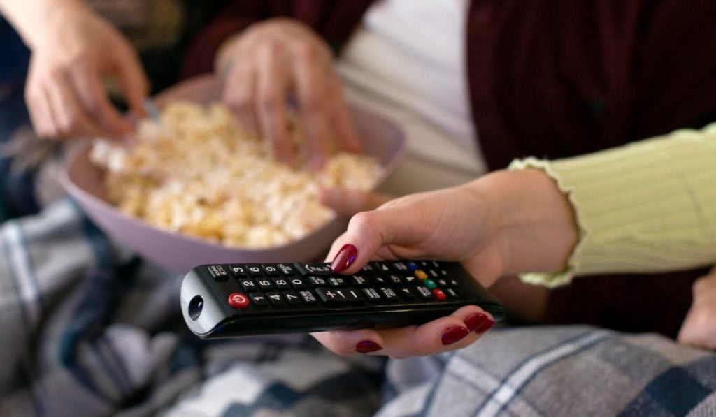 A hand with painted nails holding a Samsung remote. Another two hands grab popcorn from a bowl