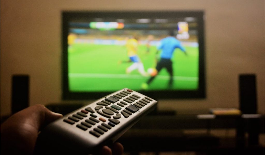 a hand holding a remote control pointing the remote at a Smart tV that is displaying a soccer match. There are two speakers, one on either side of the TV.