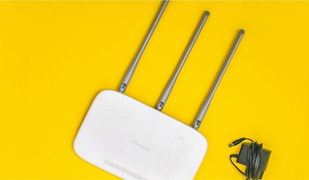 a white router with three antennas, a power adapter on yellow surface.