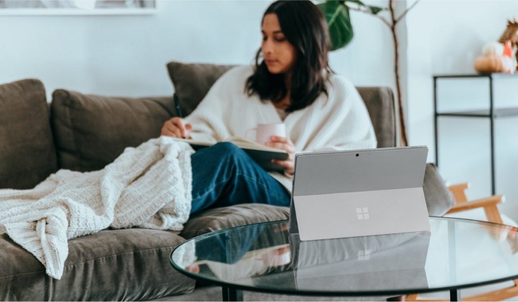 Woman laying on the couch under a white blanket. She is holding a remote control in her hand. On the coffee table next to her there is a Microsoft tablet.