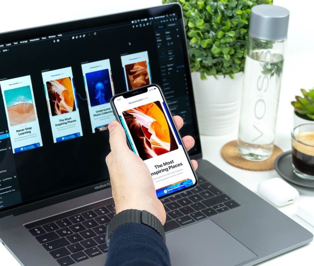 A hand holding a mobile phone in front of a laptop showing designs in Figma with water and coffee next to the computer.