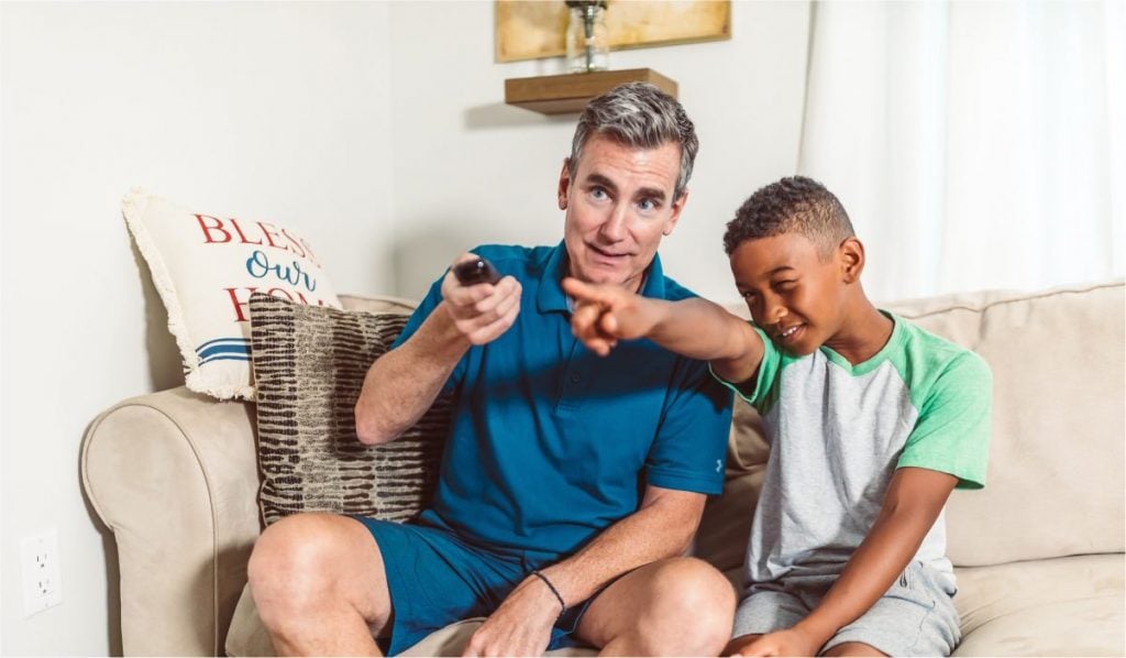 A man and a kid sit on a couch. The man is holding a remote and the kid is pointing forward.