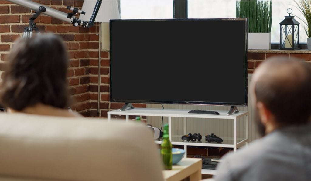 Two people sit in chairs, looking at a Smart Tv with a black scren. The Smart TV is standing on a drawer next to a brick-built wall