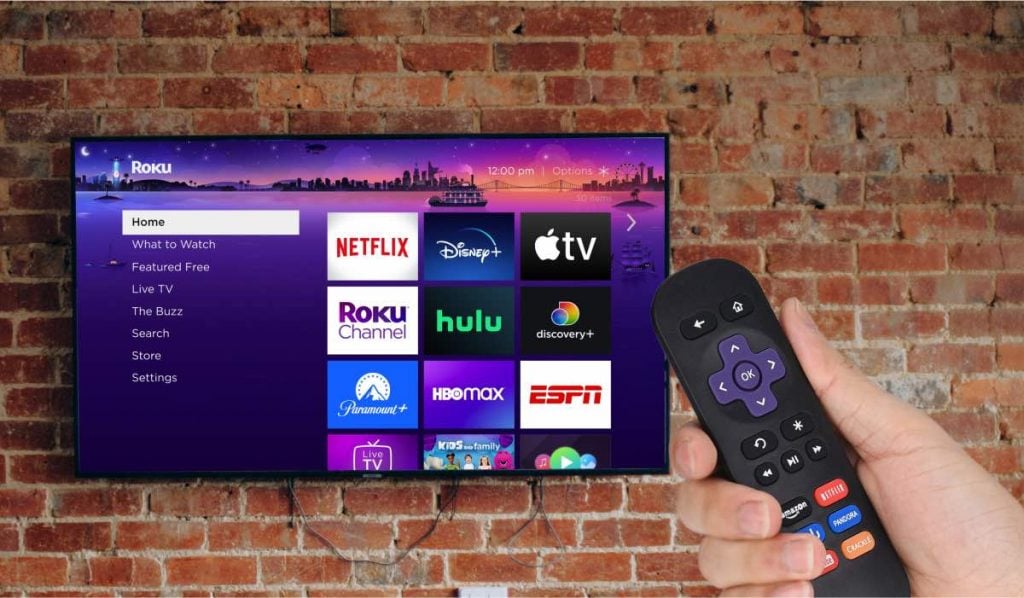 Roku interface on a TV. The TV is hanging on an exposed brick wall. There's a hand with a Roku remote, pointing at the TV