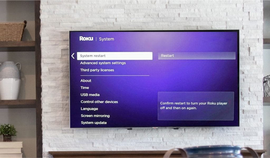 A TV is hanging on a wall in a living room. It has a Roku interface on the screenn. The highlighted part says "System Restart"