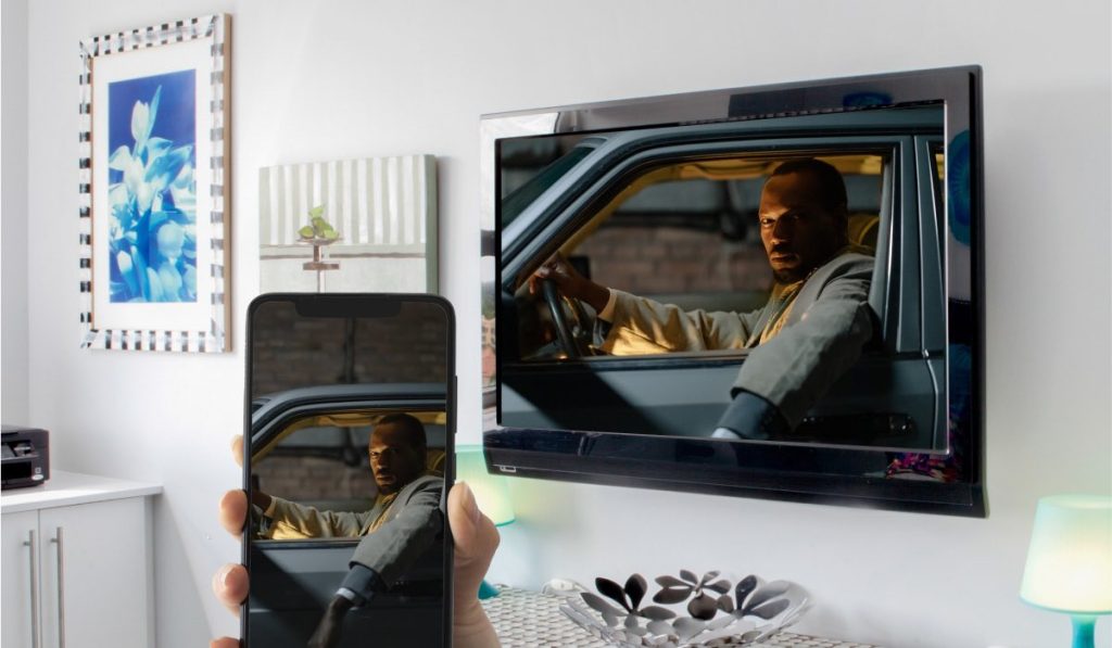 A hand holding a smartphone vertically. The smartphone is screen mirroring an image of a man sitting in a car, sticking his hand outside through an open window and looking menacingly. The image is mirrored to a wall-mounted TV on a wall with two other images.