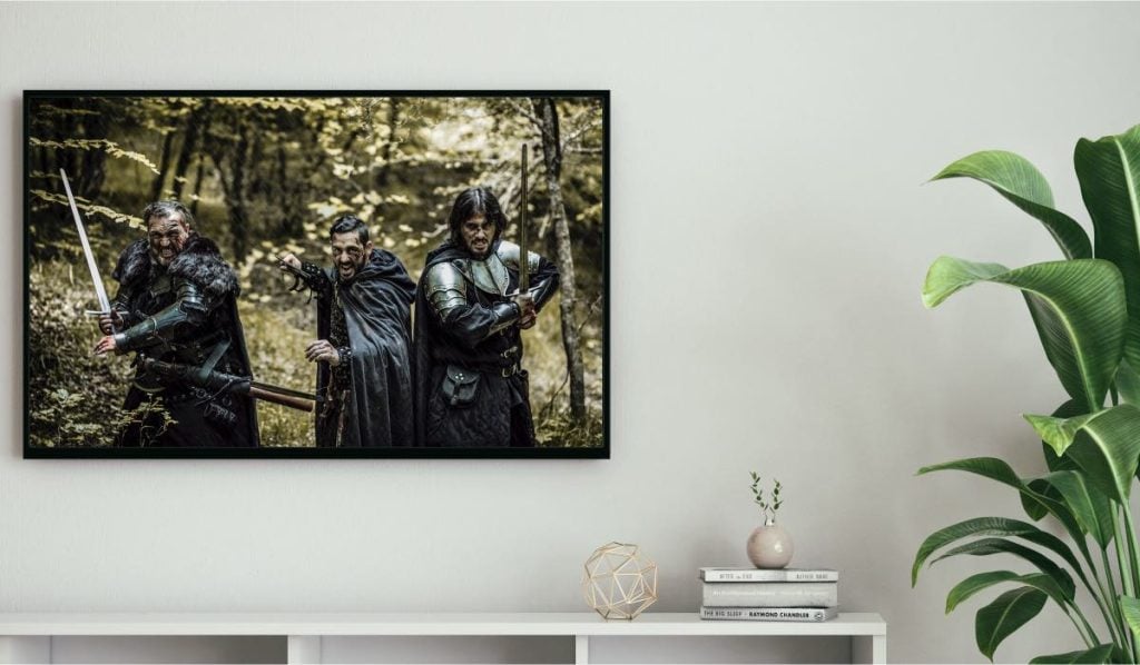 Three men in knights clothes on a TV screen. The TV is hanging on a wall, above a drawer and next to a plant