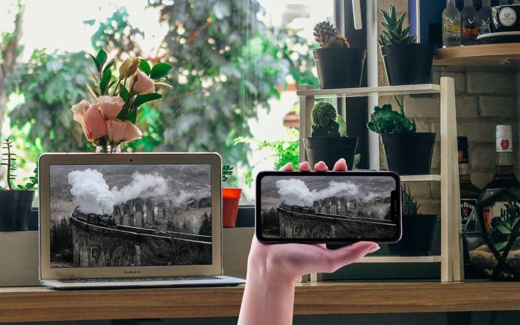A MacBook on a desk by the window, there are books on the shelves and plants outside the window. A hand is holding an iPhone displaying an image of Hogwart's Express. The MacBook is displaying the same image.