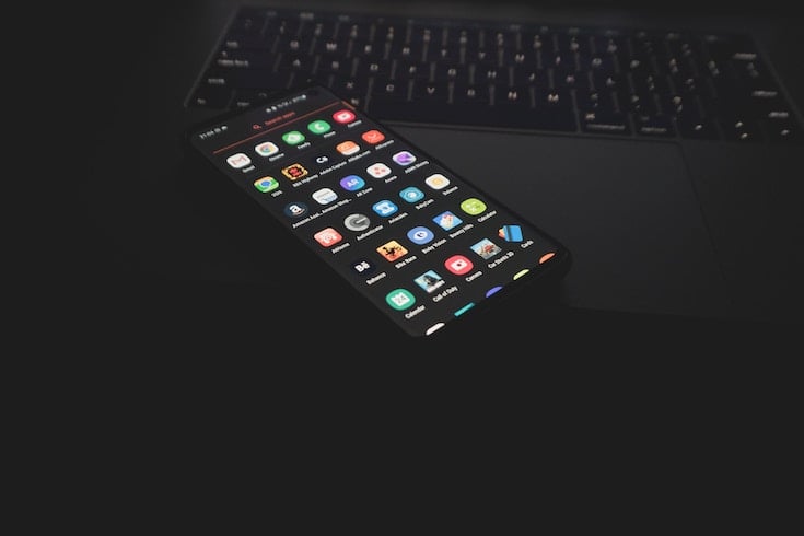 A dimmed image of an Android phone laying on a MacBook keyboard. The phone has the app menu on the screen