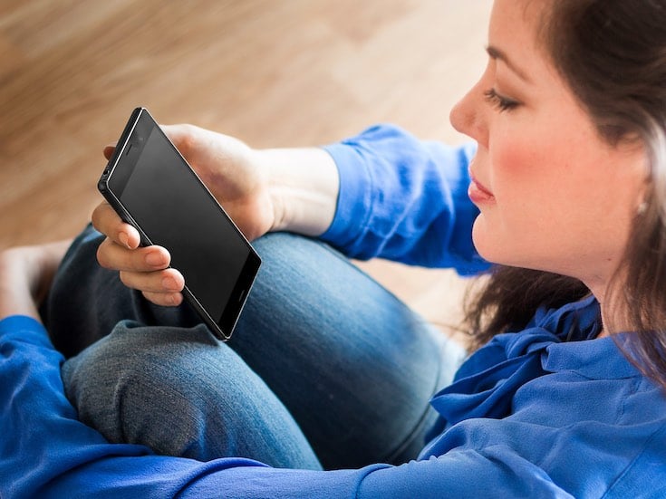 A woman sitting on the floor, holding an Android phone with a black screen.