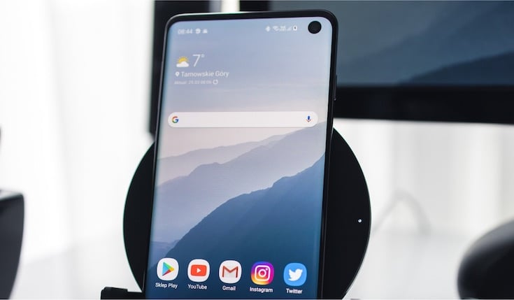 An Android smartphone with the wallpaper and several app icons on the screen. A monitor in the background
