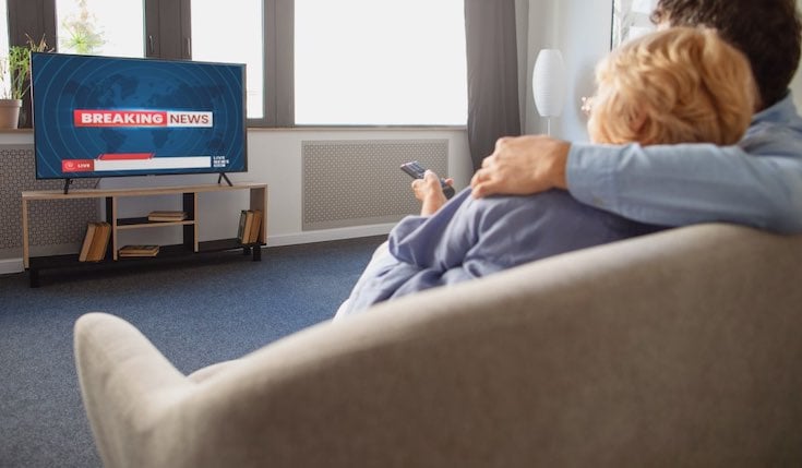 a couple in an embrace sitting on a couch. They watch 'Breaking News' on TV that is standing opposite to the couch.